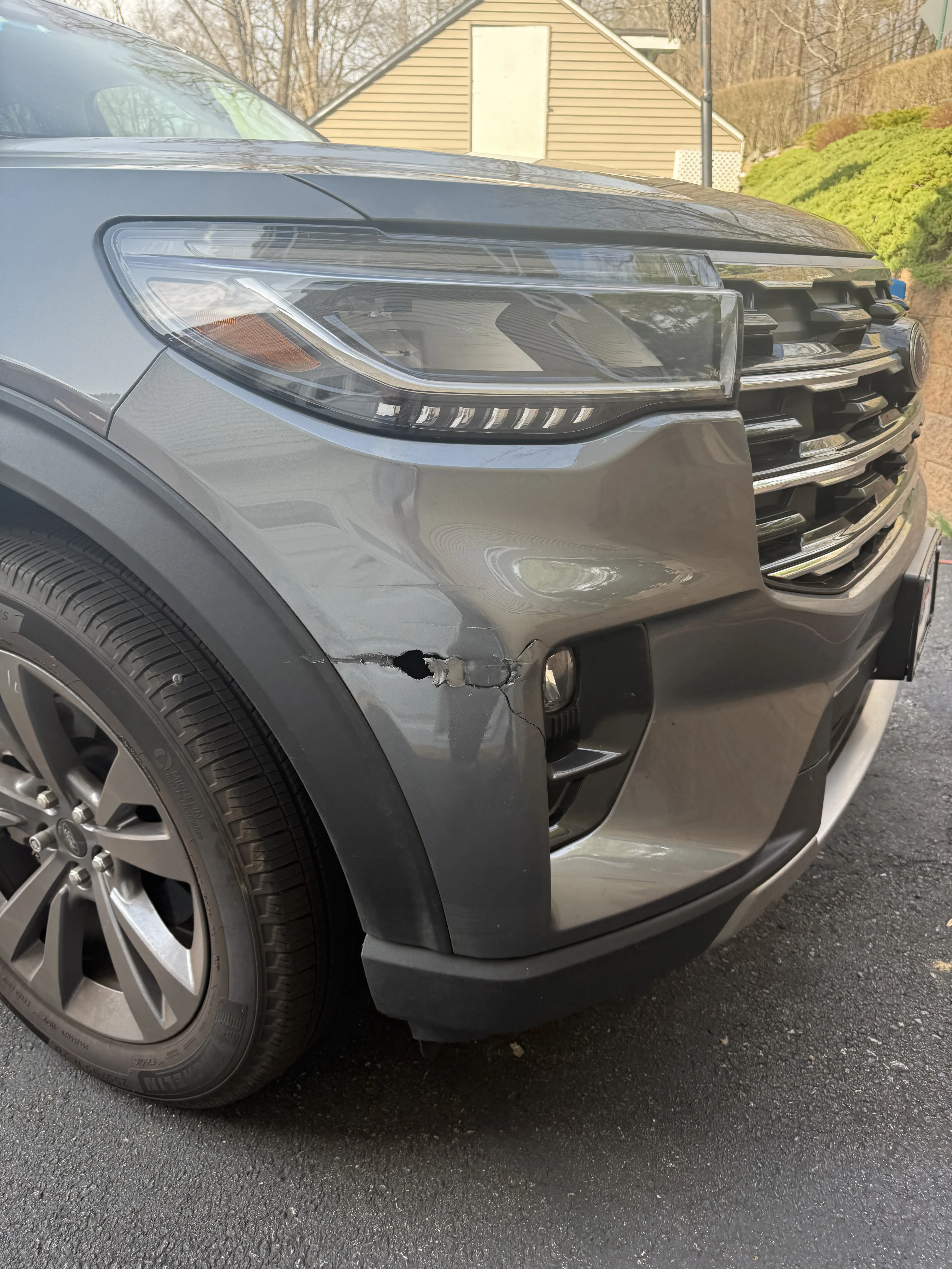 Gray Ford Explorer front bumper with visible dent and scratches on the driver side near the fog light, before repair.